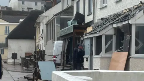 Allie Oldham A row of properties which have suffered storm damage. Tiles are falling off the roof of a porch. A man is standing in front of the properties. 
