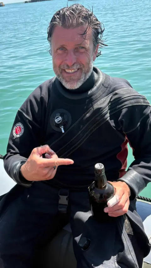 Stefan Panis in a wet suit, he is pointing at a dark glass bottle while sat on a boat on the ocean. 