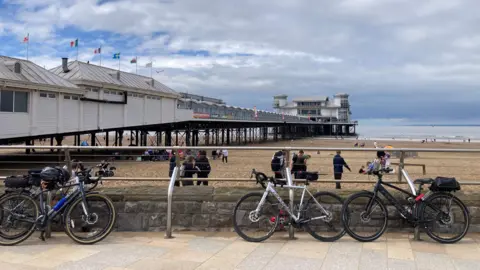 Three bicycles on stands in front of a beach and a pier