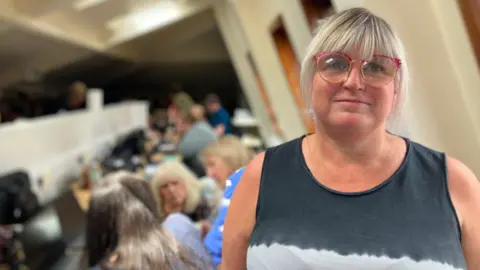 A woman with pink-rimmed glasses under a blonde fringe smiles at the camera. To the left of her, other women gather around computers.