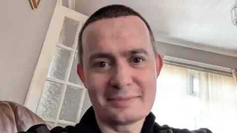 John Taylor,  with closely-cropped dark hair, is smiling at the camera as he sits in a house with and window and internal door behind him