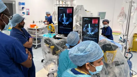 Staff wearing surgical scrubs, masks and theatre caps gather around medical equipment in an operating theatre, with imaging screens displaying procedure data.