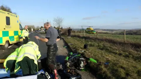 Peter Burch / Wiltshire and Bath Air Ambulance Charity The scene of a crash on a road. A significantly damaged green motorbike is lying on the ground, partially on the road and partially on a patch of grass. Two paramedics in hi-viz can be seen from behind. They are both crouched and treating someone on the ground. In the background to the left is a road ambulance and another paramedic. In a field to the right there is a yellow and green helicopter. 