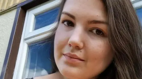 Sonia has long dark hair and brown eyes. She looks at the camera in a selfie.