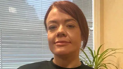 BBC Chelsea Hamilton pictured wearing a black top with her hair tied back. Her hair is brown and she has a side fringe. She is standing in front of a window with the blinds down.