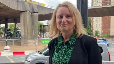 Tom MacDougall/BBC Anneliese Hutchinson has blonde hair and is wearing a black jacket and a green leafy shirt, standing next to the Park Lane roundabout - a major, traffic light controlled roundabout.