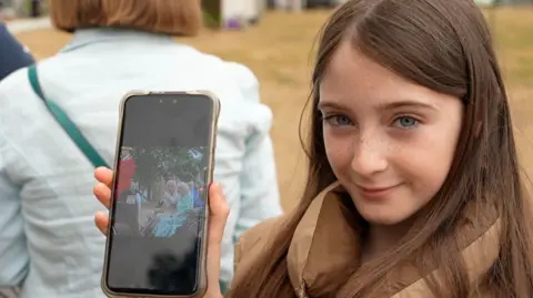 Connor Bennett/BBC Millie holds her mobile phone to reveal a photo of the King and Queen sitting in a horse-drawn carriage.