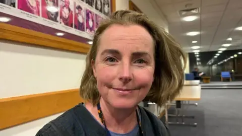 Martin Heath/BBC Lucy Bywater with medium-length brown hair, smiling at the camera wearing a grey sweater and black and orange lanyard. She is sitting in a office foyer in which a grey carpet, white wall and wooden tables are visible. There are rows of circular lights set into the ceiling.
