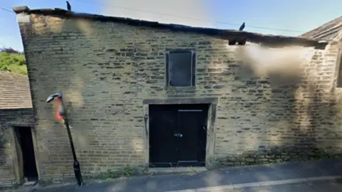 An old stone building which is blackened by soot and has several small windows which appear to be boarded up with black boards