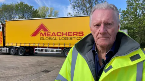 Bob Terris is an older man with white hair and is wearing a yellow hi-vis jacket. Behind him is a yellow lorry with 'Meachers Global Logistics' printed on its side.