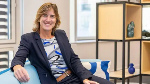 Team GB Dame Katherine Grainger sits in an office smiling at the camera. She is sat on a pale blue sofa, with her arm resting over the top of it and wears a navy blue suit, with a tan leather belt and a swirling patterned top in grey, blue and black and has dark blonde mid length hair. In the background is a wood and balck metal shelving unit which has a number of different ornaments in the shelves, they are slightly out of focus behind her.