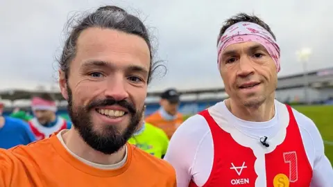 SportoGraf Two smiling runners standing side by side on an outdoor running track or field. The runner on the left has has long hair tied back and a beard. He is wearing an orange top. The runner on the right has short, dark hair and is wearing a headband and a red and white sleeveless running vest over a long‑sleeved white base layer