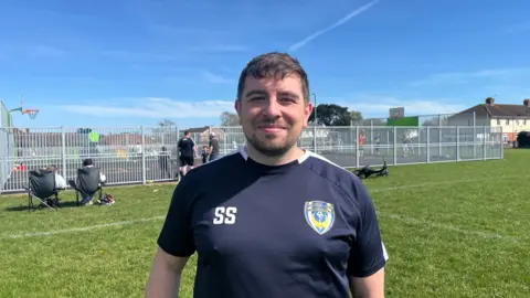 Man stood in front of big multi-use play area. He has short, black hair and is wearing a dark blue top. He has his initials on the top.