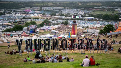 People relax on a hill near the Glastonbury sign during the Glastonbury Festival at Worthy Farm in Somerset.  In the distance there is a sea of tents that covers multiple fields. 