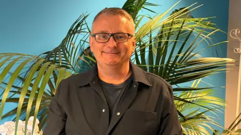 A middle-aged man in glasses stands against a blue background, with a large palm plant behind him. 