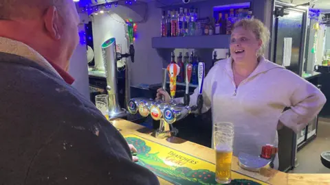 Mer Lally behind the bar at the Talbot Inn. She is smiling at a customer whose back is in vision. A half drunk pint of beer is on the bar.