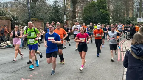 Crowds of runners work their way around the route, they are running in the road while crowds watch from the sidelines and cheer