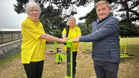 BBC Two woman wearing yellow bowel cancer guernsey shirts and a man wearing a blue puffer jacket holding onto a lime green piece of exercise equipment. They are outside and standing in front of a group of trees.