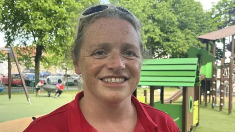 A woman with fair hair, tied back and white sunglasses on her head, is looking into the camera and smiling. It's sunny and there are green playground facilities behind her including a climbing frame and a swing. There are trees in the background.