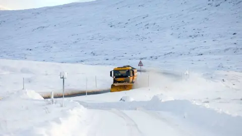PA Media A yellow gritter lorry is pictured on A87 amid a vast expanse of white snow on the road and in the hills.