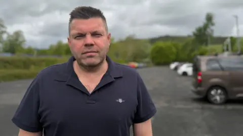 A man in a black T-Shirt with dark hair standing in what looks like a car park with trees in the background