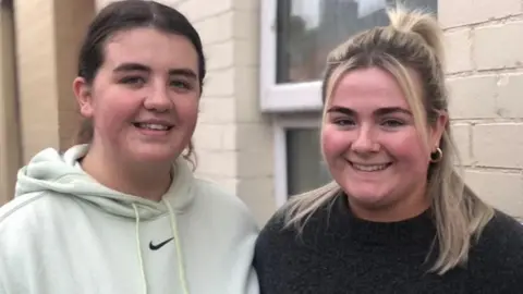 Sierra and liah outside a house with cream windows. Siara has black hair and wears a white hoodie with a nike tick. Lih has golden hair and wears a black jumper.