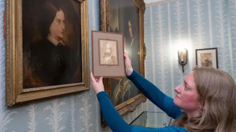 Charles Dickens Museum A female curator with light brown hair and a blue dress holds up a photograph next to a portrait on the wall of the Charles Dickens Museum, to compare the two images.