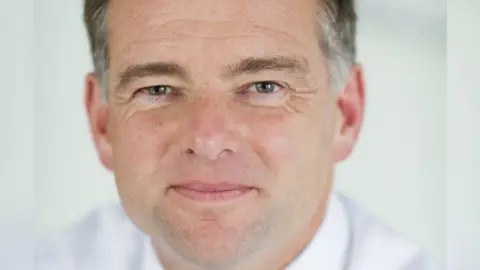 Nick Page smiles at the camera. He has grey hair and wears a white shirt.