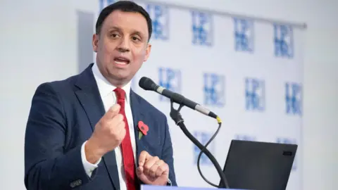 PA Media Anas Sarwar giving a speech on stage, wearing a blue suit, white shirt and red tie. He is speaking into a microphone.