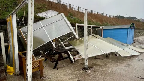 Castle Beach Cafe Screens and other beach shelter paraphernalia blown over, with a blue beach hut to the right.