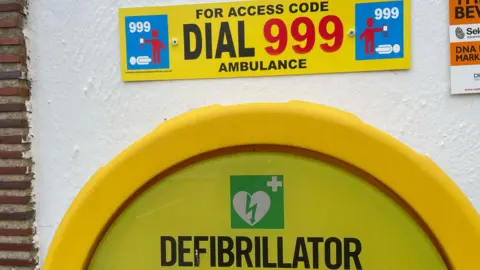 Shaun Whitmore/BBC A defibrillator cabinet with a sign saying 'for access code dial 999 ambulance'