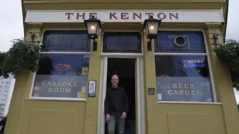 The pub is a yellowish/green colour with old fashioned signage that reads The Kenton and lamp lights. Signage in the windows reads "karaoke room" and "beer garden". A man with grey hair and wearing a black shirt and grey jeans stands in the doorway. The camera is pointing at an angle upwards from the ground. 