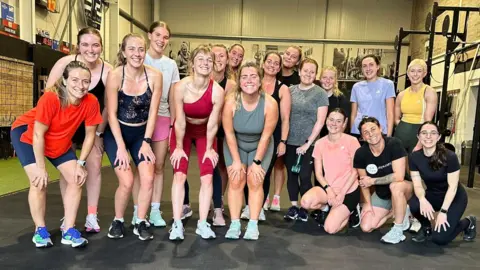 Social Girls Club A group of 17 girls wearing active-wear pose for a picture in a gym after finishing a workout.