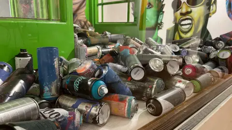 Supplied A pile of spray paint cans with a mural behind them. 