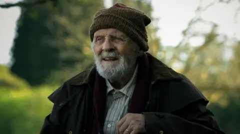 Christopher Raphael Sir Anthony Hopkins with a grey beard. He wears a dark coat, woolen hat and a beige shirt with a blue striped. 