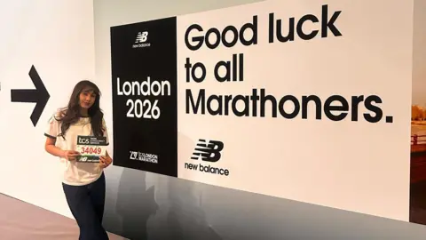 Aaliyah Jalil Aaliyah, in black bottoms, a white T-shirt, holding a marathon number badge and a large sign. She has long dark, loose hair. 