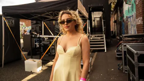 Jacinta Owen Jacinta stands backstage at a gig. She wears a beige strappy dress and black sunglasses. She has long, blonde curly hair