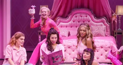 Paul Coltas The Plastics and Cady Heron sit in Regina George's bright pink bedroom and read the burn book. They're joined by Regina's mum who is sitting on the bed holding a cocktail 