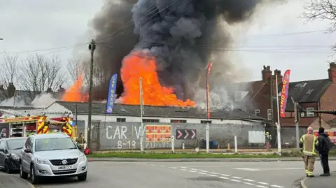 Large flames and plumes of black smoke can be seen coming from a car wash facility behind a metal fence. Emergency service workers and a fire engine are on the scene.