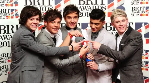 PA Archive/PA Images A group of five people in suits stand together in front of a “BRIT Awards 2012” backdrop, holding an award and pointing at it.