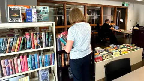 A library with many books in it. A woman with blonde bobbed hair has her back to the camera and is holding a couple of books. She is wearing a pale top and dark trousers. To the right is a horizontal book holder. To the left are shelves full of books. In the background, a man sits at a computer next to a bank of windows.