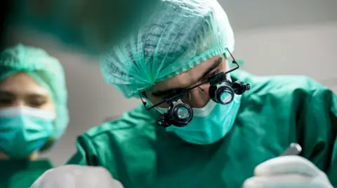 A surgeon in scrubs, leaning over someone as he performs surgery on a patient 