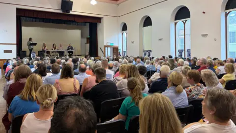 A wide shot of Coleraine Town Hall showing the room completely full of people. At the rear of the shot is the stage with a panel of speakers sitting behind a table and a woman standing at a lectern.