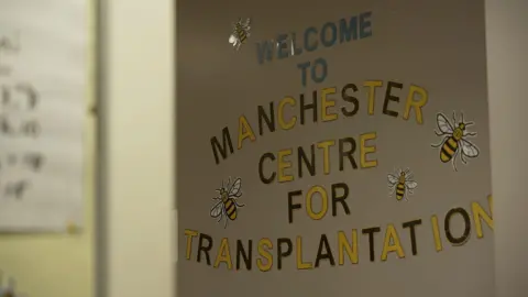 A sign on a hospital wall which reads welcome to Manchester Centre for Transplantation in yellow and black lettering