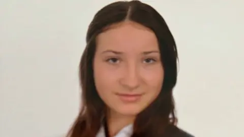 A teenage brown-haired teenage girl looks at camera through a family lecture notes by Durham police. She was wearing a white shirt, navy and white striped tie and navy blazer.