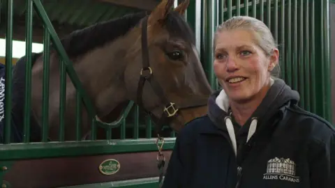 Sara stands in front of Tango the horse who is in a brown wooden stable with green metal bars. Sara wears a blue zip-up fleece, with a hoody. She is a white woman who is smiling and has blonde hair. 