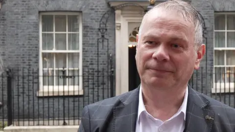 Corin Dalby, with receding grey hair and wearing a white shirt and grey suit with a lighter grey pattern, stands in the rain across the road from 10 Downing Street.
