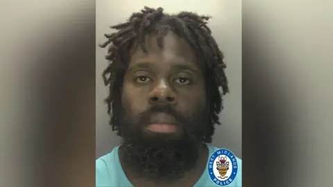 Police mugshot of a man with dreadlocks and a dark beard and moustache. he is wearing a pale blue T-shirt and facing the camera.