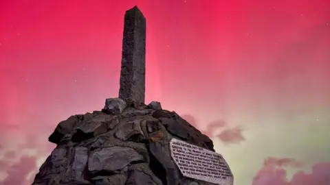 The plinth of the Guthries memorial on the Isle of Man, with the Northern Lights in the background: a bright pink sky with a  light green hue behind. Stars can be seen twinkling in the pink sky.
