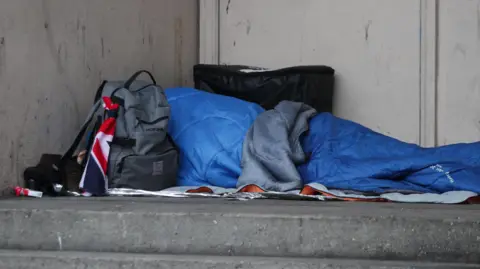 A stock image of a person sleeping rough in a doorway, they are hidden by their sleeping bag and are surrounded by their belongings in rucksacks